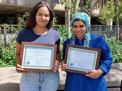 Two interns standing in front of a tree holding their certificates of completion.