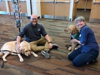 Intern and supervisor sitting on the ground with two therapy dogs. There is a rotating mental health brochure stand in the background.